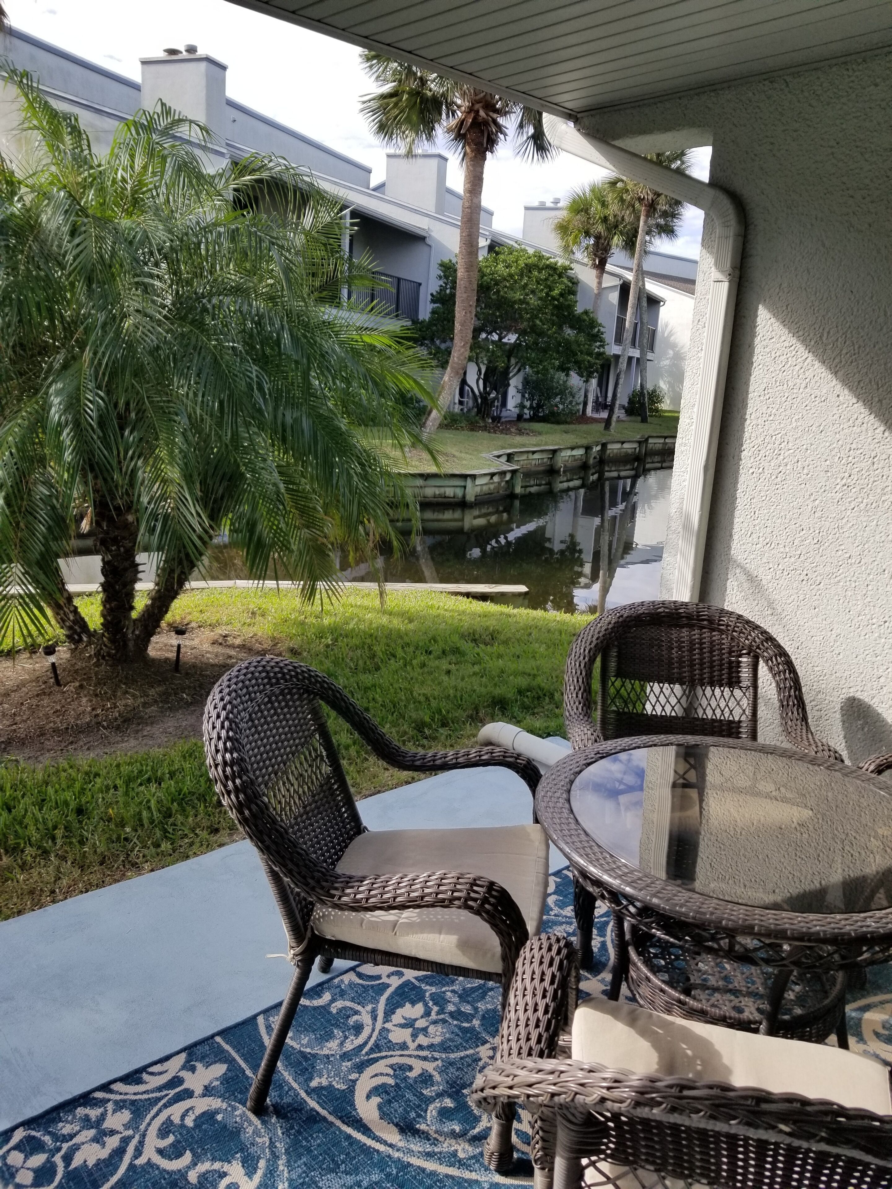A patio area with wicker furniture and a glass table, overlooking a small pond and greenery.