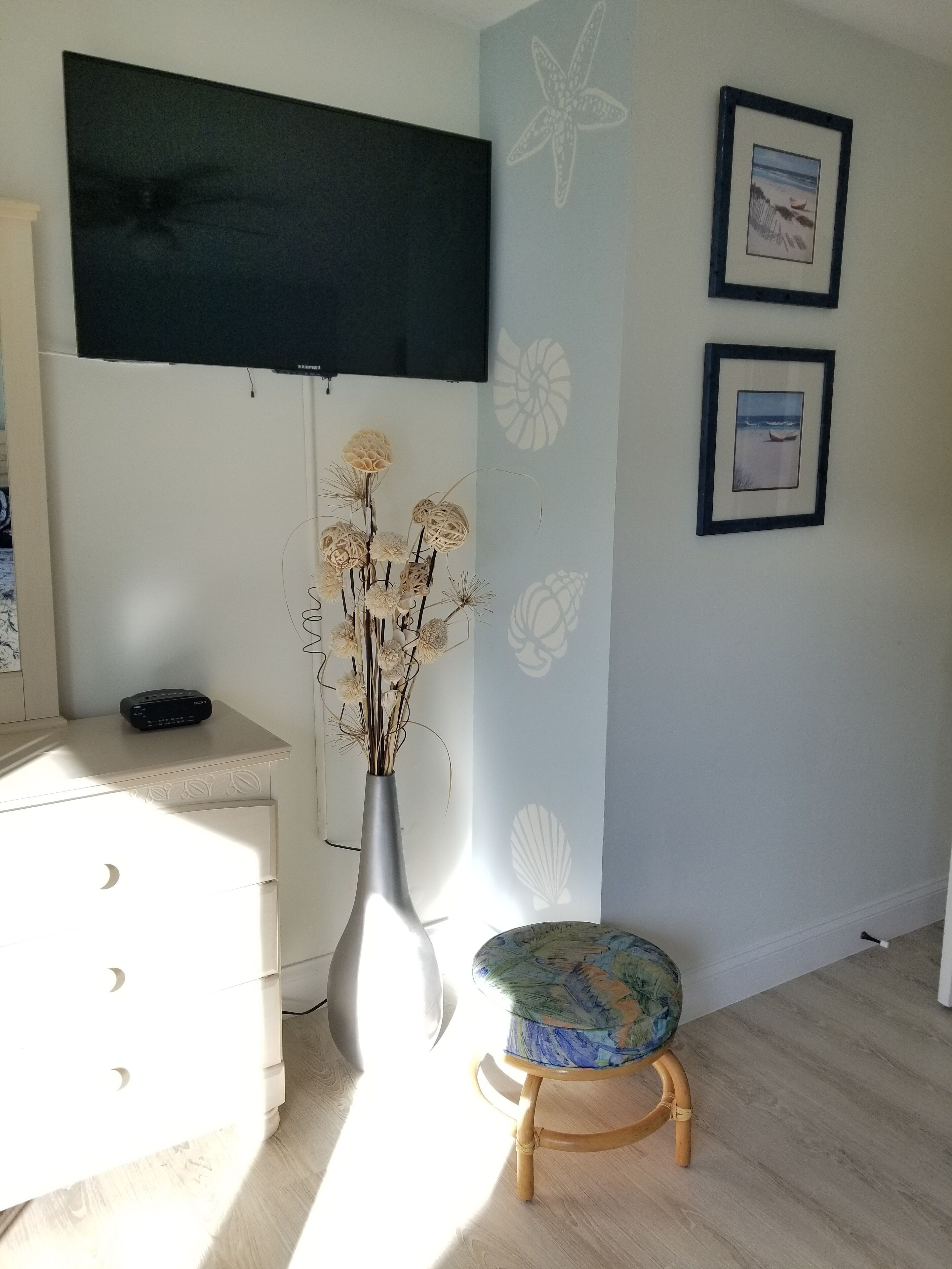 A room with a white dresser, a wooden stool, a vase with dried flowers, a flat-screen TV, and two framed pictures on the wall.