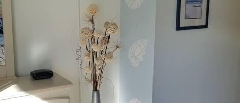 A room with a white dresser, a wooden stool, a vase with dried flowers, a flat-screen TV, and two framed pictures on the wall.