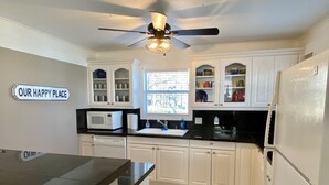 A kitchen with white cabinets, a black countertop, a ceiling fan, and a sign that reads "OUR HAPPY PLACE". - Cozy Lakefront Cottage on Eagle Mountain Lake (Azle)