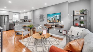 A modern living room with a grey sofa, a coffee table, and a TV. There is a kitchen in the background with white cabinets and a refrigerator. - ★ Stunning 3BR Modern Charm in South Philly ★ (Philadelphia)
