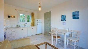 Een kleine keuken met witte kastjes, een koelkast, een eettafel met stoelen en een raam met gordijnen.