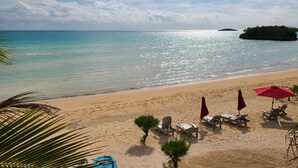 Una playa con agua turquesa cristalina, una orilla arenosa y una pequeña isla a lo lejos. Hay sillones de playa y sombrillas dispuestas para el descanso.