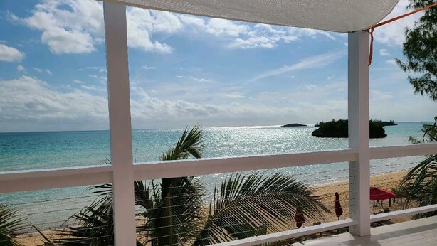 Vista de una playa desde un área techada con barandilla, palmeras y una isla distante.
