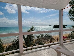 A beach view from a covered area with a railing, palm trees, and a distant island. - Trela Sands is a 3 Bedroom Unit with generator, right on a sandy beach (South Palmetto Point)