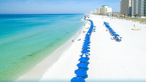 A beach with rows of blue umbrellas and white sand, with buildings in the background.