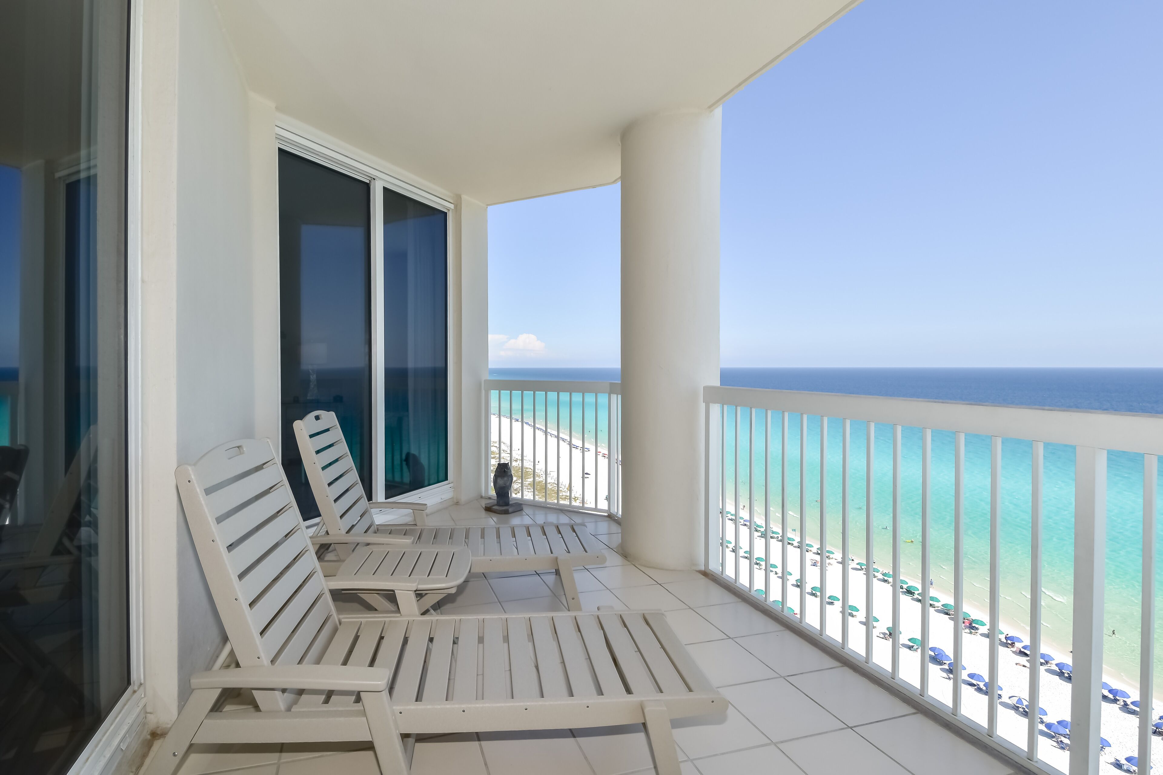 A balcony with white chairs and a table, overlooking a beach with umbrellas and a clear view of the ocean.