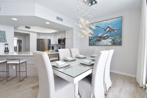 Uma área de jantar moderna com uma mesa de vidro posta para seis pessoas, cadeiras brancas e um quadro com tartarugas marinhas na parede.
