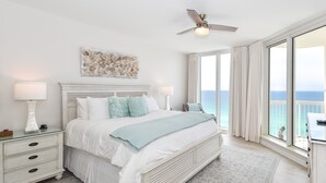 A bedroom with a large bed, a ceiling fan, a view of the ocean, and a nightstand with a lamp.