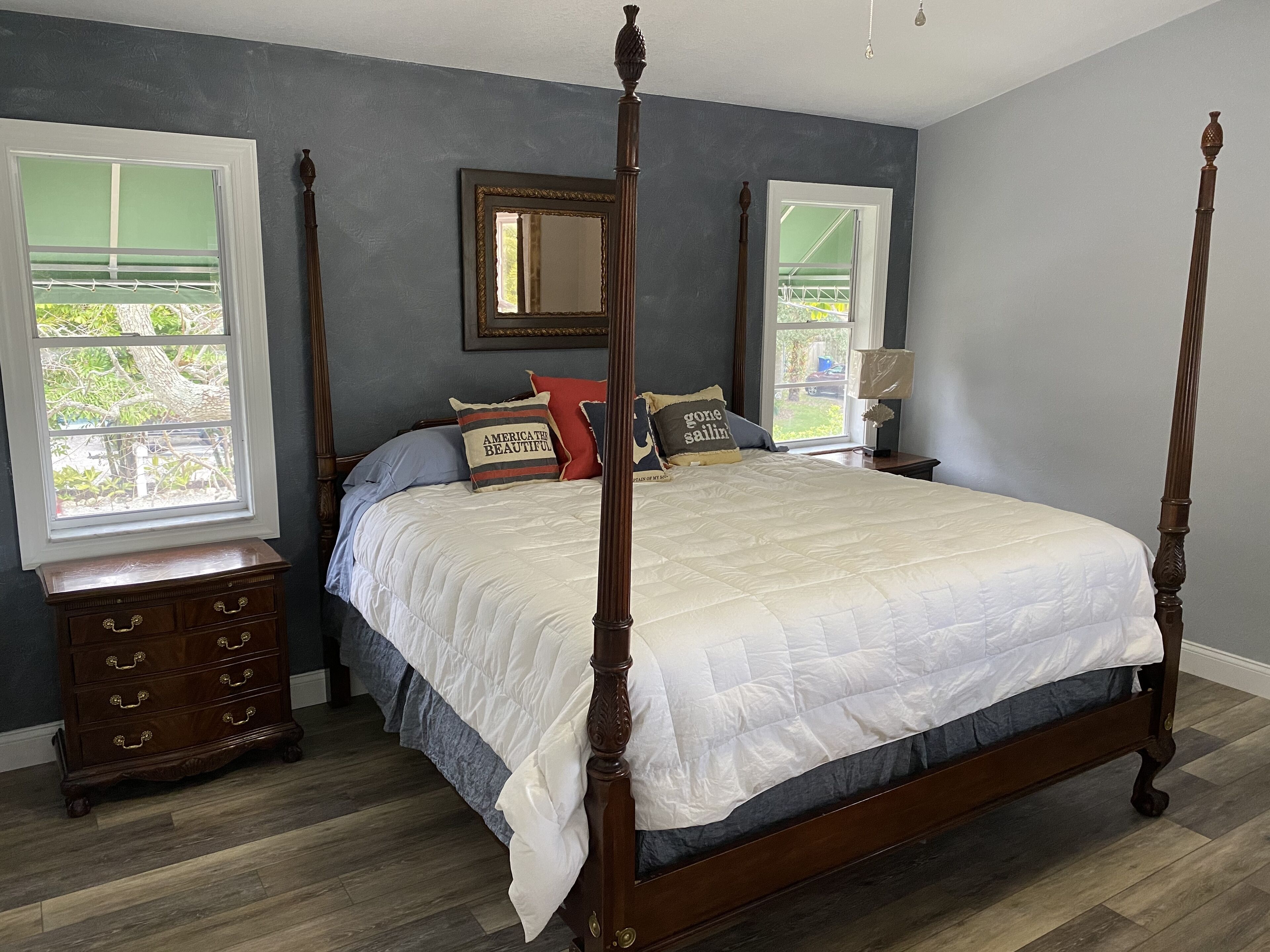 A four-poster bed with a white quilt, a dresser with drawers, a mirror, and two windows with blinds.