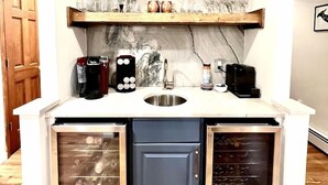 Cocina compacta con un lavamanos empotrado, dos refrigeradores pequeños, una cafetera y estantes superiores con varias botellas y vasos.