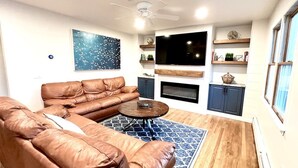 Una sala de estar con un sofá de cuero marrón, una mesa de centro redonda, un tapete azul estampado, un televisor de pantalla plana, estantes de madera y una chimenea.
