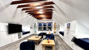 Una habitación con techo inclinado, vigas de madera, un ventanal grande, un televisor, un sofá, una mesa y una cama.