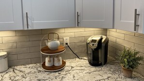 Una cucina con mobili bianchi, una macchina da caffè, un bollitore, un dispenser per zucchero e sale, e una pianta in vaso.