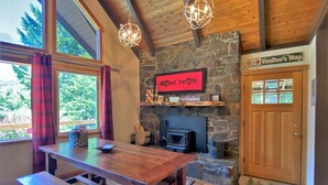 A cozy cabin interior with a stone fireplace, wooden beams, and a large window overlooking greenery. - Cottonwood Chalet (Packwood)
