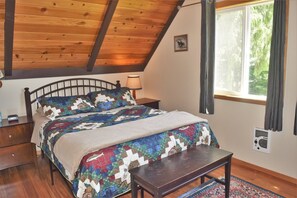 Een slaapkamer met een houten plafond, een bed met een sprei, een nachtkastje, een lamp, een raam met gordijnen en een radiator.