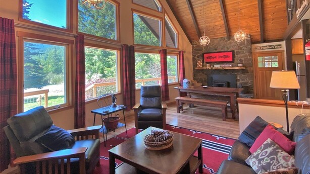 A spacious living room with large windows, a stone fireplace, and a high ceiling with wooden beams. - Cottonwood Chalet (Packwood)