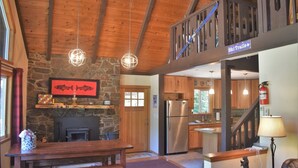 A cozy cabin interior with a stone fireplace, wooden beams, a kitchen with a stainless steel refrigerator, and a staircase leading to a loft area. - Cottonwood Chalet (Packwood)