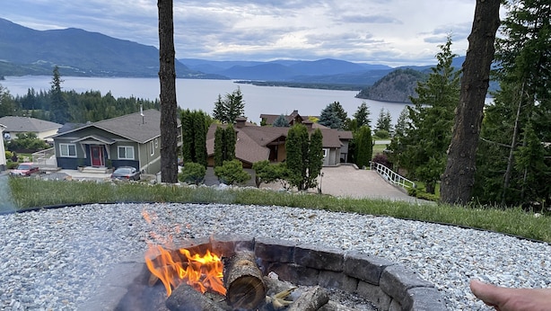 Un foyer avec des bûches et des flammes, entouré de gravier, offrant une vue sur un lac et des maisons, avec des montagnes en arrière-plan.
