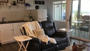 Un salon chaleureux avec un canapé en cuir noir, une petite table en bois et une vue sur l’extérieur grâce à de grandes fenêtres.