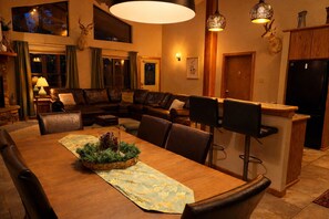 Un vaste salon comprenant une grande table à manger en bois, des canapés en cuir et un coin cuisine avec un réfrigérateur noir.