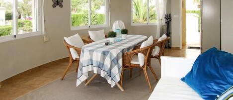 A room with a dining table, chairs, a bed, and a view of greenery outside.
