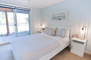 A bedroom with a large bed, a nightstand, a bedside table, a framed picture on the wall, and a sliding glass door leading to an outdoor area.