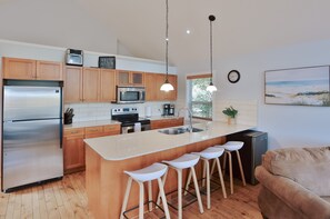 A modern kitchen with wooden cabinets, stainless steel appliances, a central island with bar stools, and a large window providing natural light.