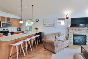 A modern kitchen with a central island, bar stools, a stone fireplace, a sofa, and a flat-screen TV.