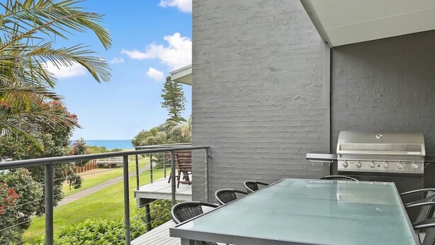 Un balcon avec une table et des chaises en verre, un barbecue et une vue sur l’océan.