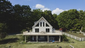 A white two-story house with a balcony, surrounded by a grassy yard and trees. - Modern 5 Acre Farm Retreat with Game Room, Views, Privacy, and Proximity! (Pine Bush)