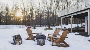 Een sneeuwbedekte buitenruimte met Adirondack-stoelen en een houtkachel, met op de achtergrond kale bomen en een gebouw met een witte gevel.