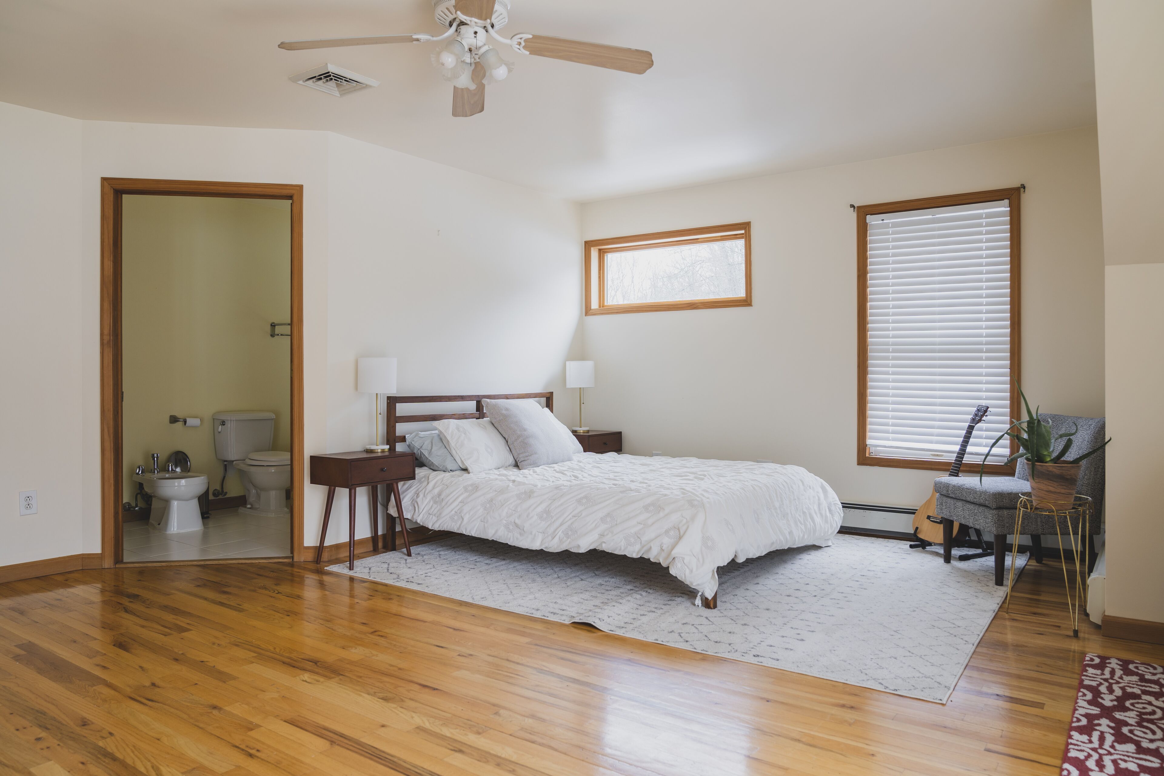 Une chambre avec un plancher en bois, un lit avec une literie blanche, une table de chevet, une lampe, une guitare et une chaise avec une plante.