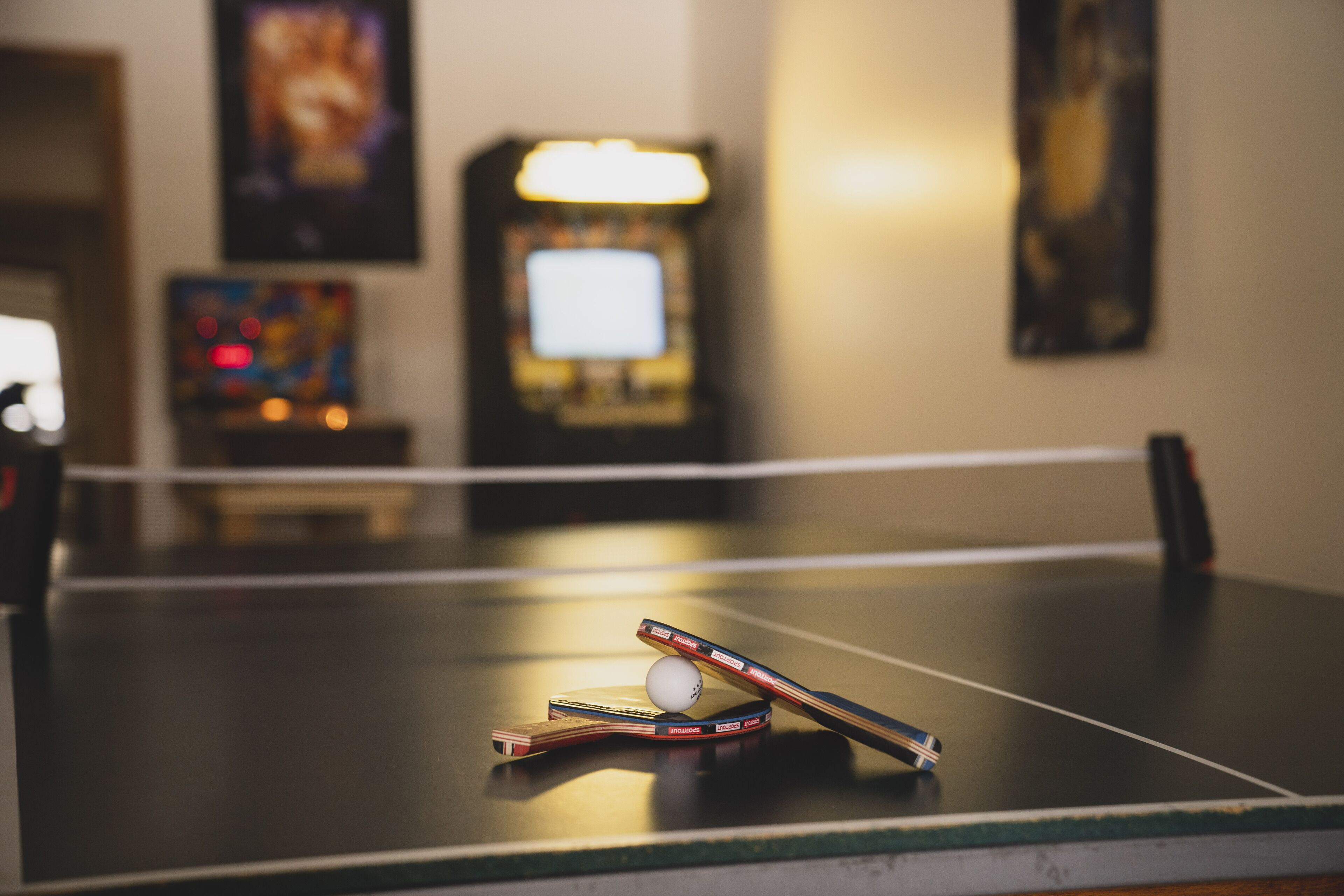 Una mesa de ping pong con una pelota y paletas en primer plano, una pantalla de televisión al fondo y máquinas recreativas.