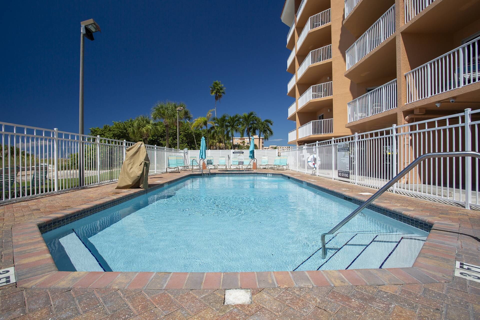 A swimming pool with a tiled deck, surrounded by a white fence, and adjacent to a multi-story building with balconies.