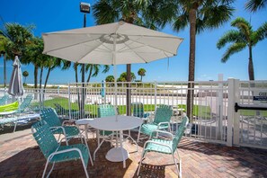 A patio with a table and chairs, a white umbrella, and a view of the beach and palm trees. - Beachfront Condo. Heated Pool. Activities, Fun, End Unit, Parking, & Great Location! (Treasure Island)