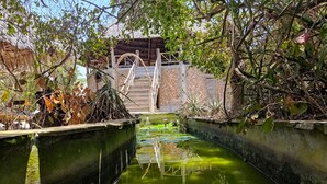 A narrow water channel with a wooden bridge, surrounded by vegetation and a building with a thatched roof in the background.