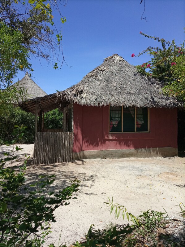 Une hutte à toit de chaume, aux murs peints en rouge, entourée d’une clôture en bois et posée sur un sol sablonneux.