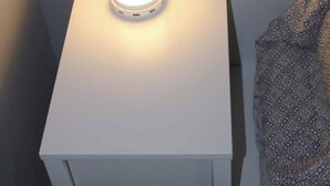 Una mesita de noche blanca con una lámpara, una manta estampada en la cama y una pared lisa.