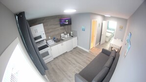 Un espacio habitable compacto con kitchenette, un sofá gris y un televisor montado en la pared.