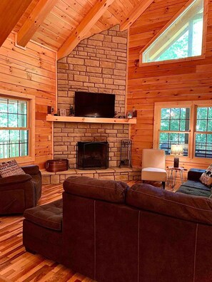 A cozy cabin interior with a stone fireplace, a television mounted above it, and a comfortable seating area with a sofa and armchairs. - NEWER cabin, 5 BR, 2 fireplaces, hot tub, privacy (Logan)