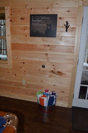A wooden cabin interior with a wall-mounted sign, a metal bucket with colorful items, and a door to the right. - NEWER cabin, 5 BR, 2 fireplaces, hot tub, privacy (Logan)