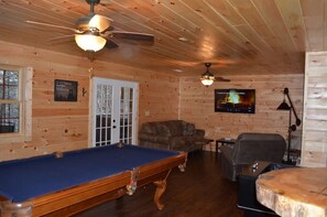 A cozy cabin interior with a pool table, a sofa, a television, and a ceiling fan.