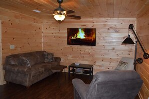 A cozy room with a wooden interior, a leather sofa, a grey armchair, a small table, a television displaying a fire, and a ceiling fan. - NEWER cabin, 5 BR, 2 fireplaces, hot tub, privacy (Logan)