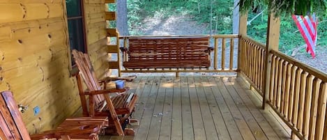 Una cabaña de madera con un porche, sillas y un banco de madera, y vista a un área boscosa.