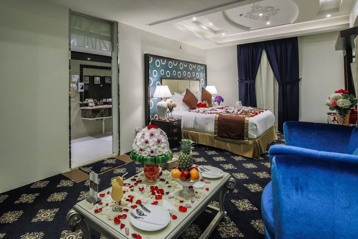 A hotel room with a neatly made bed, a dining table set for two, and a blue armchair.