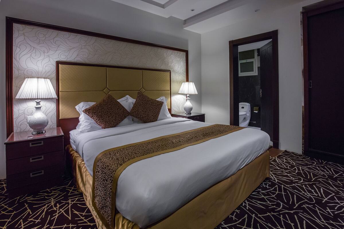A hotel room with a large bed, two bedside tables with lamps, a wooden headboard, and a patterned carpet.