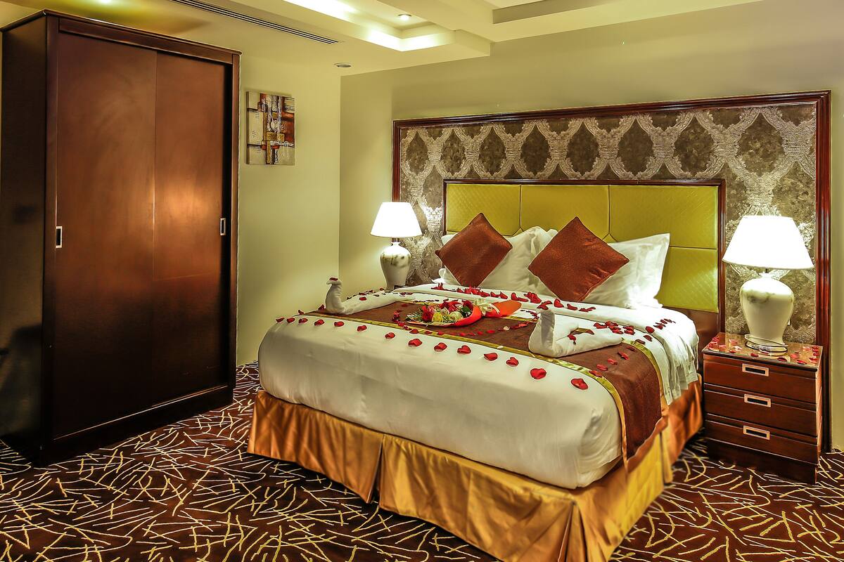A hotel room with a large bed, two bedside tables with lamps, a wardrobe, and a decorative ceiling.