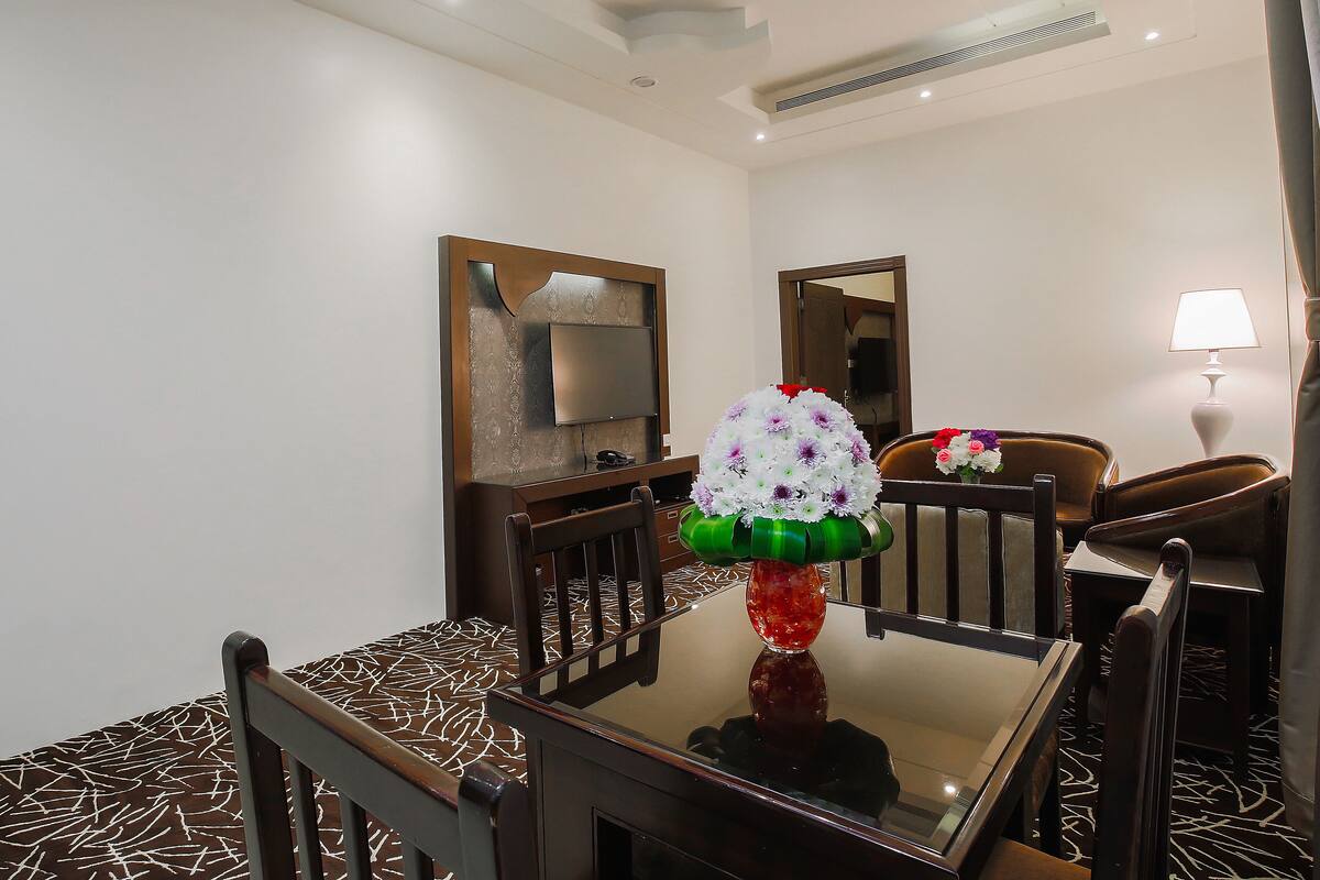 A hotel room with a dining table, chairs, a TV mounted on the wall, a mirror, a lamp, and a vase with flowers.