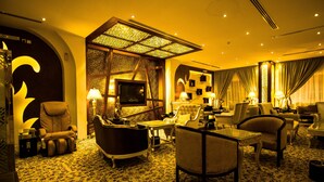 A luxurious hotel lobby with a classic design, featuring a grand piano, plush seating, and a large screen.